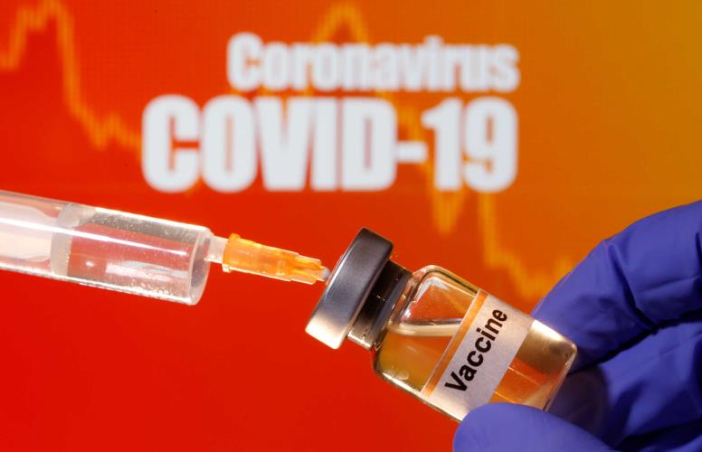 A small bottle labeled with a "Vaccine" sticker is held near a medical syringe in front of displayed "Coronavirus COVID-19" words in this illustration